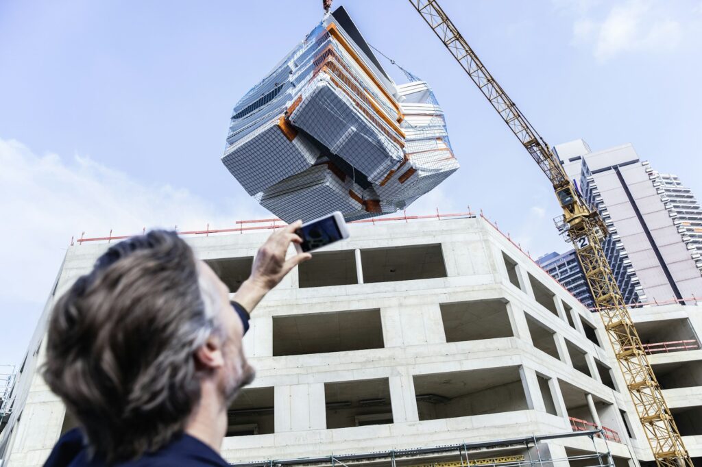 Modular construction: Man takes a picture of a modular building unit being lifted by a crane at a construction site. Prefabricated building.